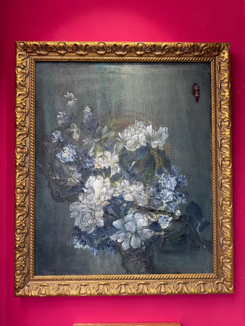 A framed still life painting of white and blue flowers on a dark gray-blue background, enclosed in an ornate gold frame and mounted against a bright pink wall.